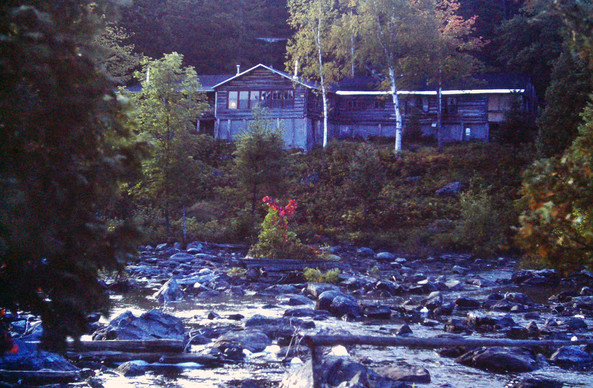 Early evening in the forest. A wide, shallow stream churns over and around dozens of gray boulders in its rocky bed. A large, rustic wooden building stands atop a small, scrubby hill rising from the stream bank. The surrounding forest is barely beginning to turn color, although a scattering of reds and oranges can be found in the underbrush.