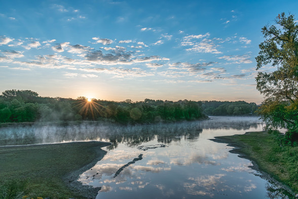 Image of the morning sun just rising above the treeline on the horizon with a creek and river in the foreground and a blue sky filled with white and grey clouds above.