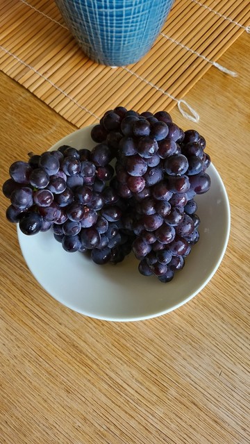 2 densely pack purple bunches of red grapes sit in a white ceramic bowl on a wooden kitchen table.