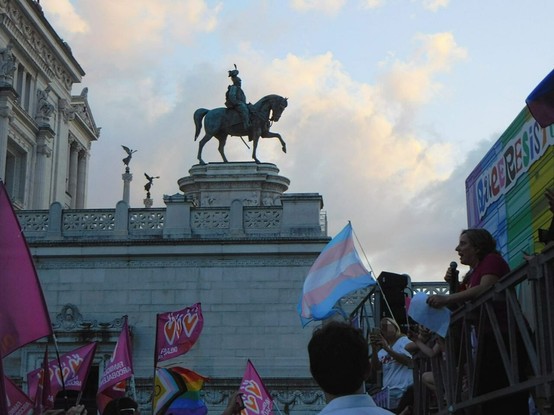 official Rome  gay pride rally .    many pride flags in the bottom side of the picture   ( a  trans one,  the progress pride rainbow one  and   many other pink ones ,  which belong to an association called  'Famiglie Arcobaleno')     and also  a stage  on the right .     a woman is taking a speech  on it .       in the background ,   an equestrian statue  ( detail  of the  'Milite Ignoto'  building in  Piazza Venezia,  which is also  the classic  final destination of Rome Pride)   and the sunset