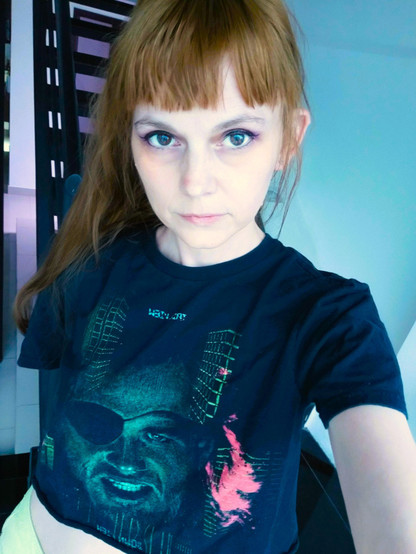 Me, a brunette woman, wearing a crop t-shirt featuring the face of Snake Plissken from Escape from New York, as well as some of the iconic green building wireframes. I also have neon green jorts on but they are mostly out of frame. My eye makeup is actually pretty good for once!