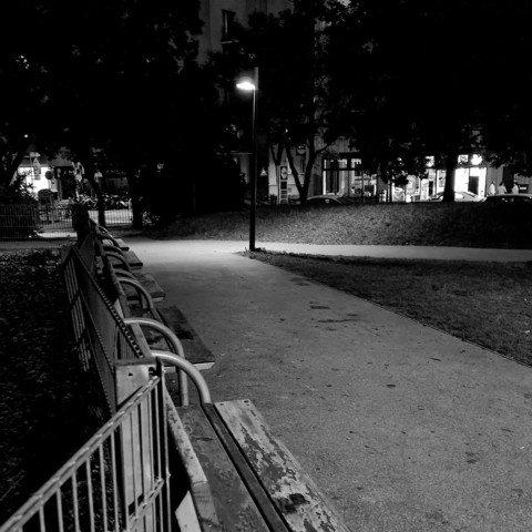 A deserted row of benches next to a footpath illuminated by a streetlight. A few lighted apartment buildings are visible behind trees.