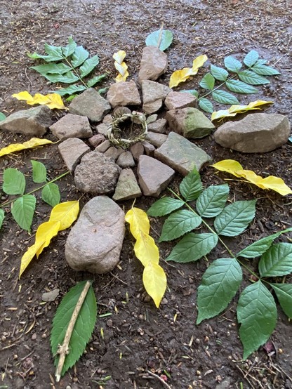 A small nature altar. A tiny circular wreath make of yew in the centre. Around it is a ring of small stones, with another ring of larger stones around that. Then there are rays, like the sun, coming out from the centre made of alternating large green leaves, more stones, and rows of bright yellow leaves.