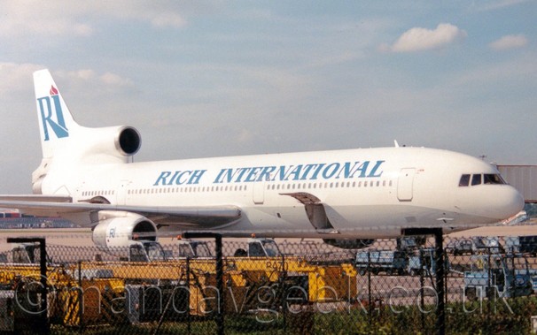 Side view of a 3 engined jet airliner parked facing to the left, with an airbridge attached to the forward door on the opposite side.
The plane is mostly white, with a silver belly, and blue "RICH INTERNATIONAL" titles on the upper forward fuselage.
The tail has large blue letters "RI", with a red flame coming out of the top of the "I".
This same logo appears in a smaller form on the engine pods under the wing.
In the foreground, 2 rows of black chainlink fence is strung across the lower 1/3rd of the frame, supported by black metal frames, and topped with razorwire, beyond which some trucks with long, extendable yellow booms with large yellow buckets on the end are lined up side by side.
Pale blue sky filled with smears of cloud fill the rest of the frame.