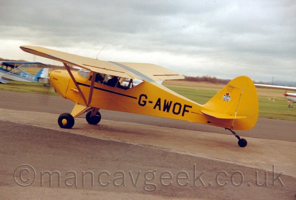 Side view of a high-winged, single engined light aircraft with a tailwheel insead of a nosewheel taxiing from right to left on a concrete taxiway.
The plane is a bright yellow, with a black stripe running along the body, and the registration "G-AWOF" on the rear fuselage in black.
In the background, a couple of other light planes are visible on the left and right of the frame, while the grass airfield stretches off into the distance, with trees marking it's edges.
The sky above it all is a dull grey with white patches.
