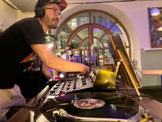 DJ behind the turntables in a warmly lit vinyl bar, arched wooden panorama window behind.