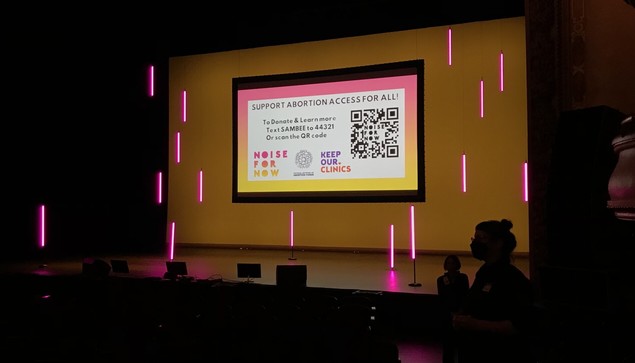 Stage with pink neon lighting and a slide "Support Abortion Access for All, To Donate & Learn more text SAMBEE to 44321"
