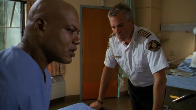 Teal'c sitzt als Patient auf einem Krankenhausbett.

O'Neill lehnt sich daran mit beiden Armen an.

Er trägt ein Feuerwehrhemd und schaut etwas zerknirscht.