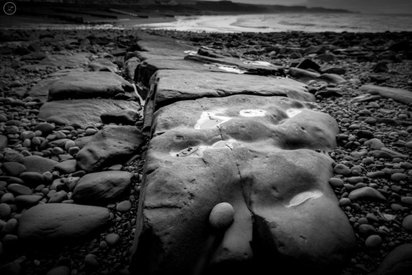 Monochrome shot of wave smoothed rock line on beach. Sea in background and pebble beach to left and right of frame
