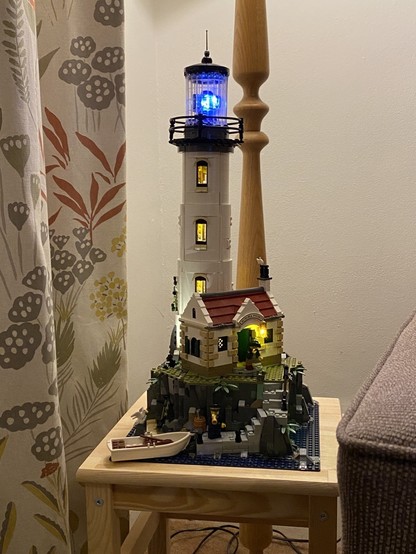 The Lego motorised lighthouse set sitting on a table in a corner of a room. Additional lights have been fitted into each floor of the tower, and a very bright LED has been fitted to the rotating beam light. A flickering lantern hangs by the entrance to the lighthouse keepers cottage.