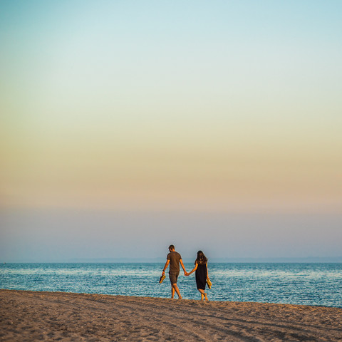 An einem Strand laufen im warmen Licht der untergehenden Sonne zwei Menschen Hand in Hand dem Meer entgegen. Man sieht sie von hinten und wÃ¼rde sie als Mann und Frau lesen. Beide tragen kurze, gedeckt einfarbige Kleidung und halten ihre Schuhe in der jeweils Ã¤uÃŸeren Hand. Das Foto ist quadratisch, der Horizont liegt am unteren Viertel. Ãœber den Personen ist sehr viel unstrukturierter, aber von Blau- Ã¼ber Orange- in LilatÃ¶ne verlaufender Himmel.