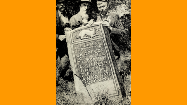 Photograph of an old gravestone being held up by three men – above the inscription is a carving of an animal with a spear through it.