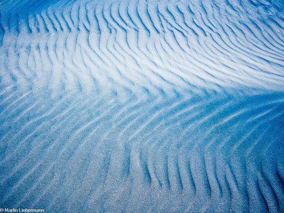 Photography of a sand dune at dusk. Small ripples patterns in the sand, caused by wind, are appear as softly curving lines in a vertical direction. A larger, but horizontal  sand wave causes a  distortion in  the smaller ripples. 

The shadows of the white sand are tinted blue from sky at dusk, the lights are faintly purple from the remainnig colors of sunset. Single grains of sand are visible, making the image appear soft.