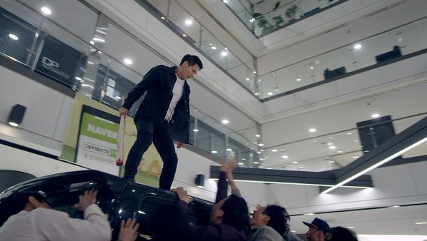 A still from the film Gangnam Zombie. A young Korean man stands on top of a car in a shopping mall, surrounded by zombies who are reaching up towards him. Image courtesy of WellGoUSA.