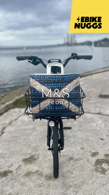 A bike, front-on, in front of the river Tay. Thereâ€™s a paper bag in the front basket with a Scotland flag design picked out in food.