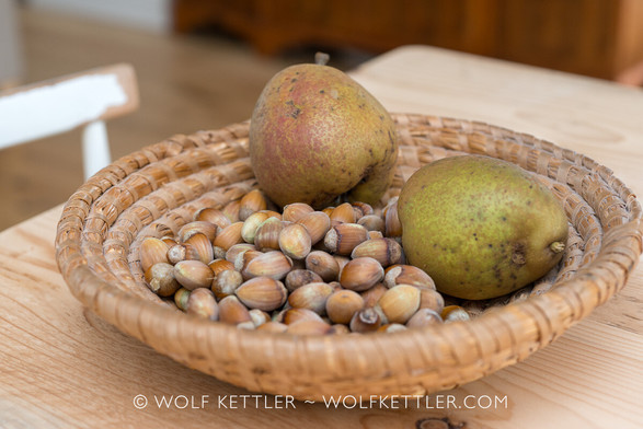 Photograph shows a rustic woven bowl containing nuts and two pears, all freshly harvested. The bowl stands on a table with a wooden top. Viewing angle is from slightly above. To the left the indication of the backrest of a white painted, worn, wooden chair. In the background the feet and bottom of a larger cabinet made from red-brown wood, standing on a light wooden floor. Chair and cabinet blurred.