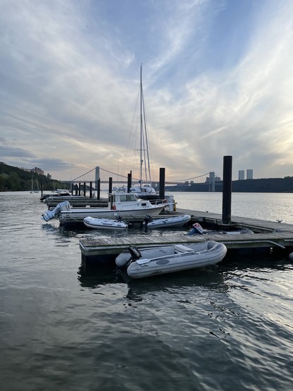 Looking south down the Hudson river towards the George Washington Bridge. In the foreground is a marina with zodiacs, a catamaran and keelboats moored in the distance. The clouds tinged red by the setting sun.