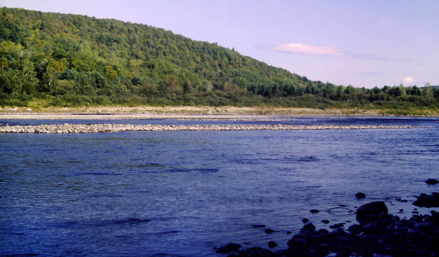 Beneath a hazy blue sky with a few streaky clouds we are standing at on a shaded rocky bank of a wide but shallow river. The opposite shore is also a jumble of rocks and small boulders, a long, narrow bar of ricks and gravel lies not far from that riverbank. A low wooded ridge rises above the far bank.