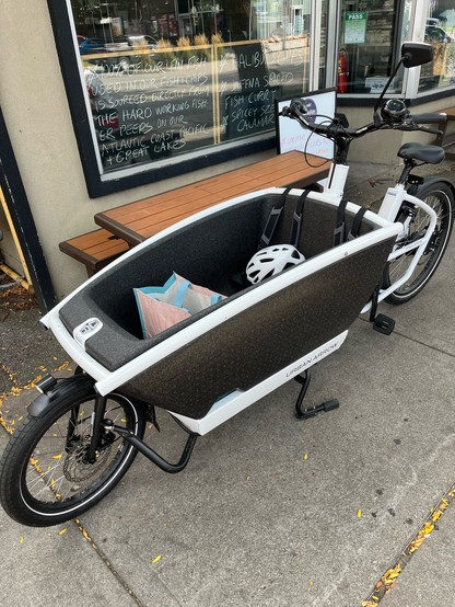 Picture of a white Urban Arrow, a family e-bike with a carrier for one adult or up to three small children. The bike is parked outside of a restaurant, and a shopping bag and a helmet are in the carrier while the riders dine.