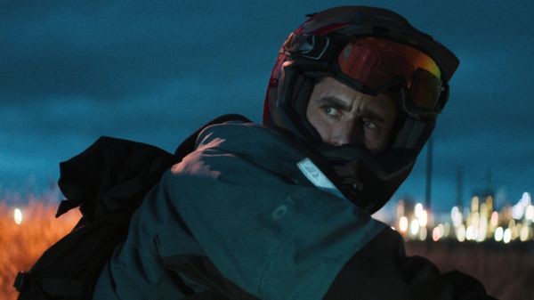 A still from the film Jackdaw. A white man in motorcycle gear looks behind him as he rides through the outskirts of a city at night.