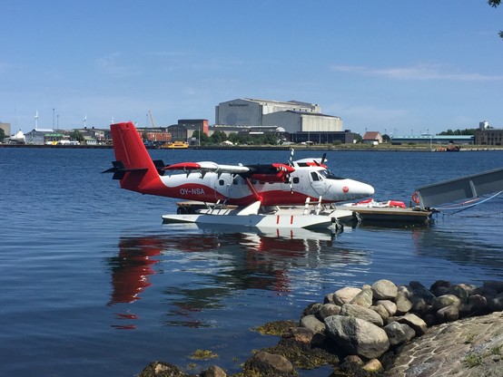 A red and white seaplane docked along a rocky coast with water shimmering underneath its floats.