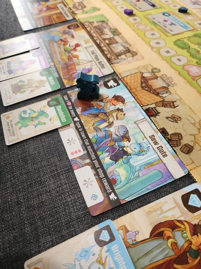 Photo of part of boardgame flamecraft. Photo of the shop sew cute which has dragons trying on clothes. Blue dragon meeple on shop
