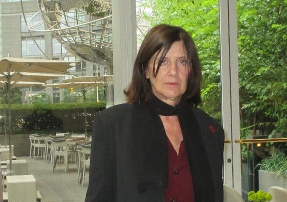 A smartly dressed middle aged white woman stands with her back to glass doors, through which an unoccupied outdoor café is visible, surrounded by trees and hedges and with a large urban building behind it. Image by Anne-Katrin Titze.