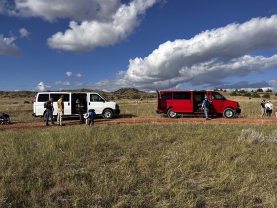 2 15-passenger vans, one white and one red, and several people preparing for fossil collecting, in a flat grassy area with a blue sky and fluffy clouds.