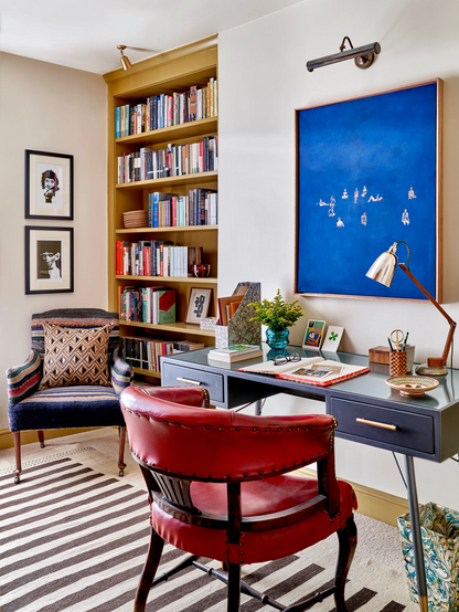 A beautiful office space in a home, with table, a bookshelf and two chairs and wall paintings.