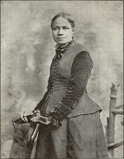 A black and white picture of Frances Ellen Watkins Harper. She stands for her portrait, gently resting her hands on the back of a chair. She looks right at the viewer and wears her hair pulled back. She has on a dark colored dress.