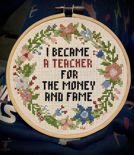Finished Crosstitch project in small hoop. It reads “I BECAME A TEACHER FOR THE MONEY AND FAME” and is circled by pink and blue flowers