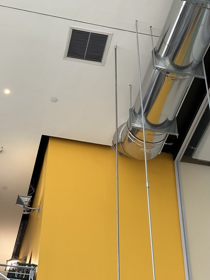 View of the ceiling with steel rods suspending a large kitchen hood underneath (off camera). A yellow wall appears adjacent to the white ceiling and a polished metal enclosure around ventilation ductwork hanging from the ceiling.