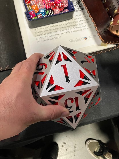 A hand grabs an 8-inch wide aluminum 20-sided die with inlaid red and black decorative resin.