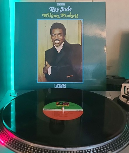 A black vinyl record sits on a turntable. Behind the turntable, a vinyl album outer sleeve is displayed. The front cover shows Wilson Pickett with arms crossed as he smiles and looks at the camera.
