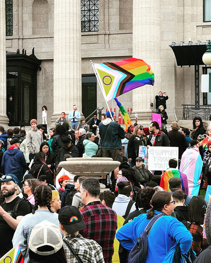 People gathered at Manitoba legislature for a protect trans youth rights rally. There is a large LGBTQIA+ flag in the centre.