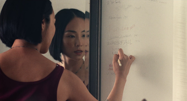 Na Young/Nora (Greta Lee) writes her name on the wall of an artist-in-residence retreat, her face is reflected in a mirror.