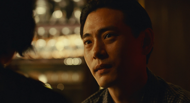 Hae Sung (Teo Yoo) listens intently to Nora, lights reflecting in his eyes.