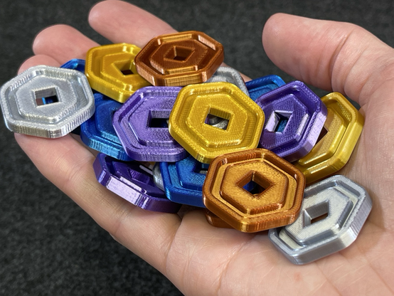 A hand holding a pile of 3d printed Robux coins from the Roblox video games, in gold, silver, copper, purple, and blue color filament