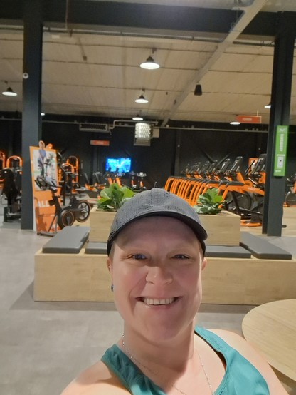 Me sitting in the gym, changing my shoes (not in the picture). I smiling at the camera, wearing a green top and black cap. Behind me some benches a f behind those, loads of orange/grey/black machines. Those are the colors of the gym.