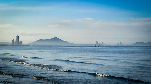 A sea view with coastline in the distance, with hills in the back country and condos along the shoreline. It is early morning, there is a low mist with blue skies above a calm sea.