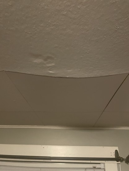 A view of a white ceiling tile that looks bowed . There are some depressions in the tile where I has pressed it with my fingers when it was wet. It is now dry but permanently deformed. It is sagging enough that there is a shadow on the wall in the background.