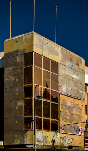 A very run-down modern narrow corner building in brutalism style with three flag poles on the roof. The central part of the building corner is made up of large orange tinted glass panes the size of the concrete slabs the building is assembled out of. The other half of the long side of the building is taken up by a large frame for a billboard of which only a couple remnants in the corner can be seen. On the glass panes one can vaguely see the silhouette of a baroque style building with a domes roof.