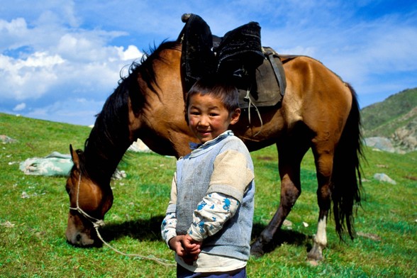 7-8yo kind standing in front of a grazing horse. Green grass and blue sky with some white clouds in a sunny day.