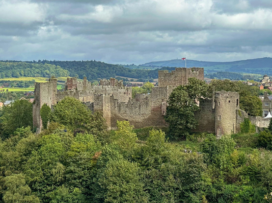 A sandy coloured old castle turrets surrounded by large mature trees and shrubs overlooking the market town of Ludlow. An English flag flies on the top of the Talleyâ€™s turret and the landscape is surrounded by several green hills and countryside.