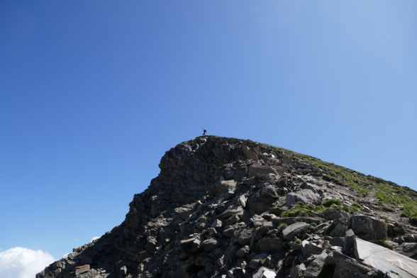 A hiker in a boasting position on the summit, seen from below. The blue sky behind him, makes him very visible, although he is very small.