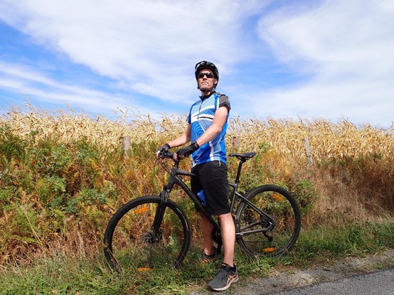 Guy in blue cycling top standing with bike in front of corn field and blue sky