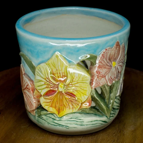 a cylindrical ceramic orchid pot, featuring releif carving of various orchid plants in flower. this view features a yellow vanta orchid veined in orange. the sky is a translucent blue. The ground is an unfortunate pale teal colour.