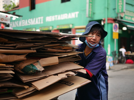 A hat wearing Chinese woman pushing stack of folded paper carton boxes on a trolley.