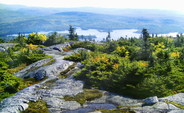 We are atop a mountain, looking out over a series of striated gray granite outcrops along the crest, partially covered by a forest of stunted dwarf spruce and fir sprinkled with a few hardwoods covered with bright golden autumn leaves. We are looking down at a huge valley dominated by a very large lake reflecting the very hazy very pale blue sky. In the misty distance beyond the lake a series of forested low mountain ranges cascade to the horizon.