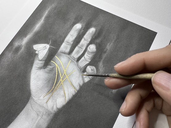 Hand with a paintbrush hovering over an art print of Chiromancy: A hand palm up, the lines in the hand painted gold. A white moths rests on the thumb. The original is graphite and gold pigments. Prints comes in two sizes.